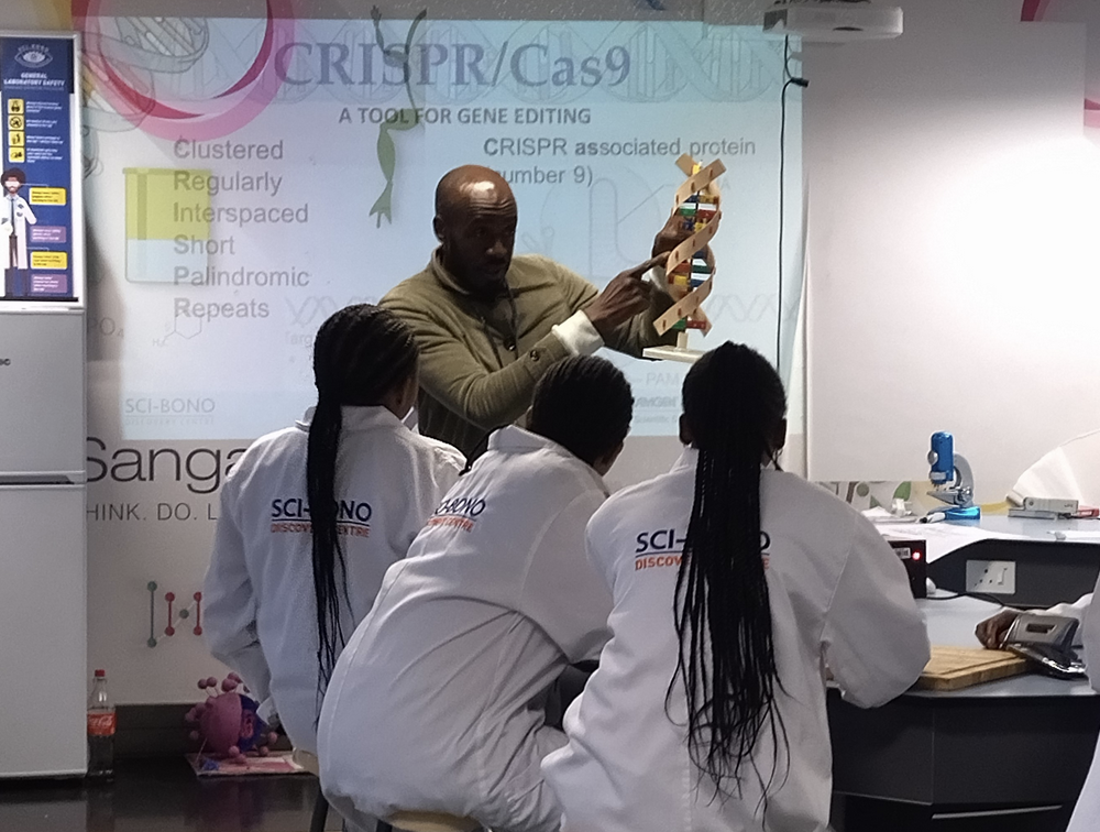 Mr Nyoni presents a model of DNA to students in labcoats. Behind him is a projection of a CRISPR/Cas9 powerpoint.