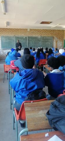 Mr Nyoni stands in front of a chalkboard and addresses a class of seated students in blue uniforms at Chipa-Tabane Secondary School.