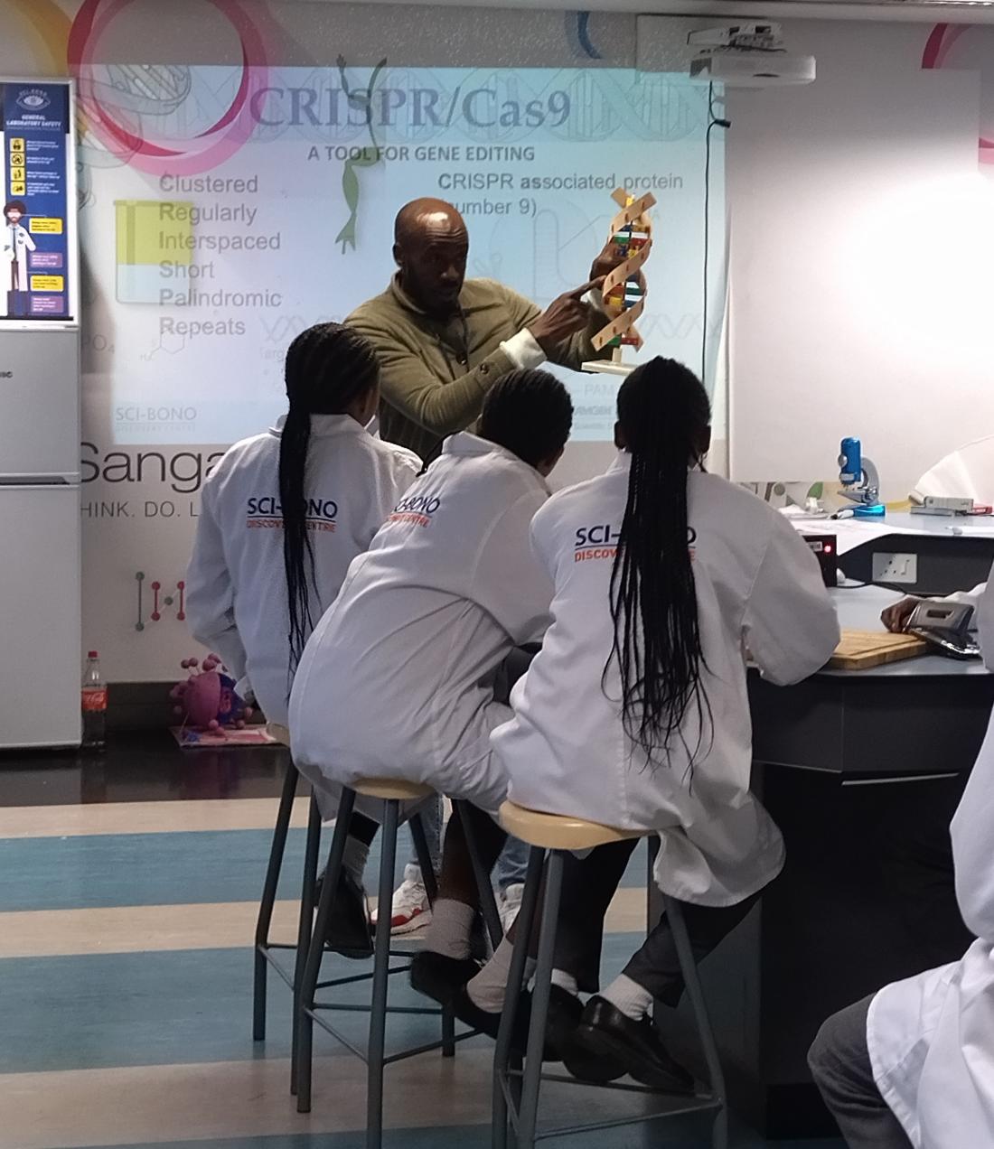 Mr Nyoni presents a model of DNA to students in labcoats. Behind him is a projection of a CRISPR/Cas9 powerpoint.